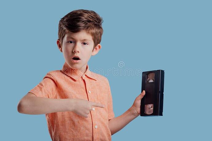 surprised-boy-showing-video-cassette-blue-background-amazed-stylish-little-kid-orange-shirt-pointing-videotape-184307497